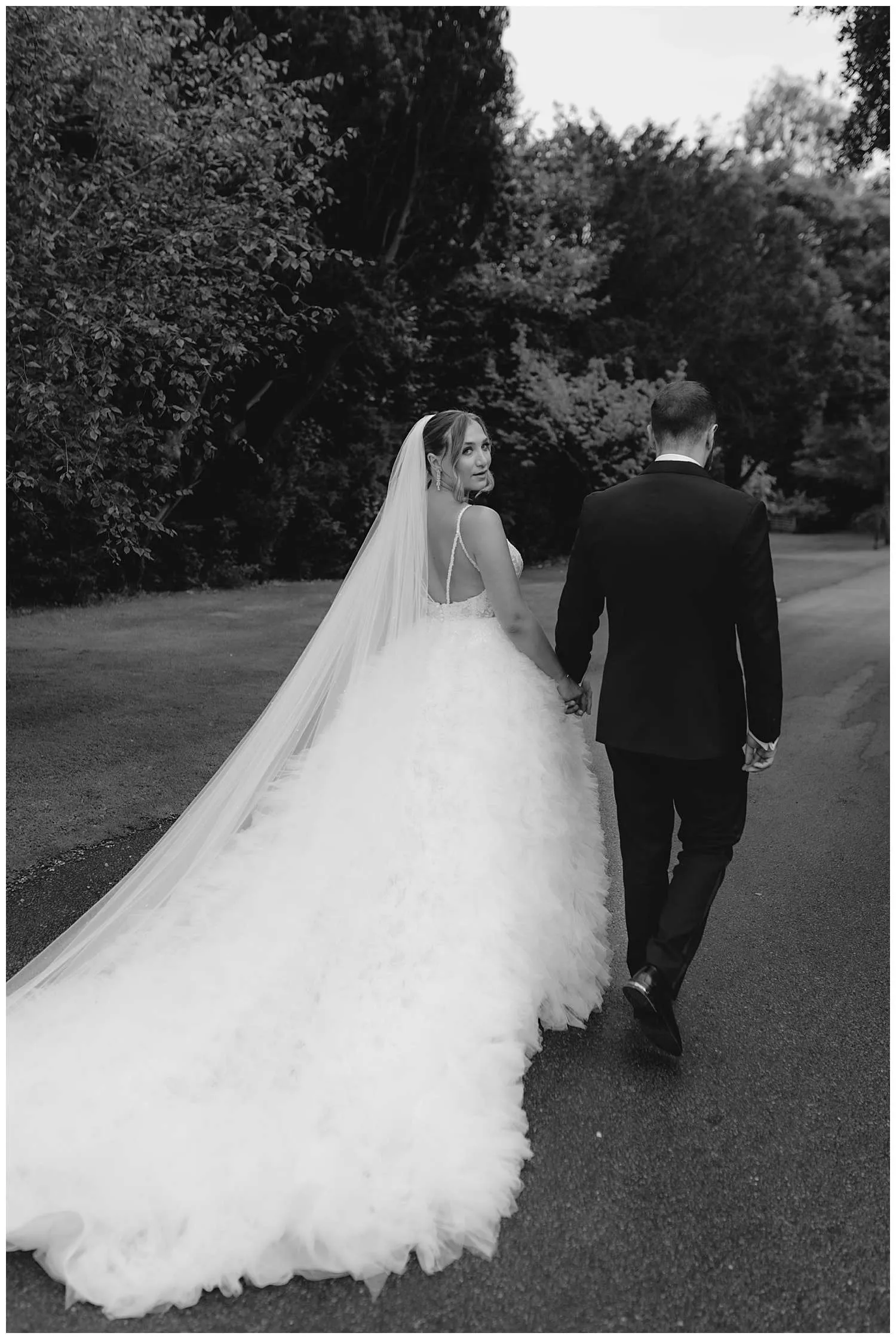 Hedingham Castle Wedding Photographer - the bride looks back towards the camera as the couple walk down the path